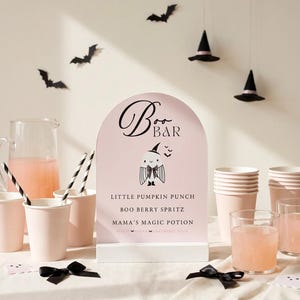 May include: An arched pink sign with a white base featuring the text Boo Bar, a ghost illustration, and a drink menu including Little Pumpkin Punch, Boo Berry Spritz, and Mama's Magic Potion. The sign is surrounded by pink cups, a pitcher, and black bows.