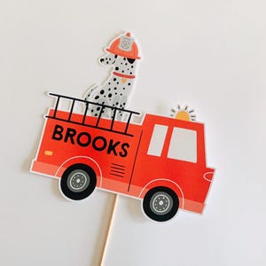 May include: A red fire engine with the name "BROOKS" printed on the side. A Dalmatian dog wearing a firefighter's helmet is on top. The truck has black wheels and a ladder.
