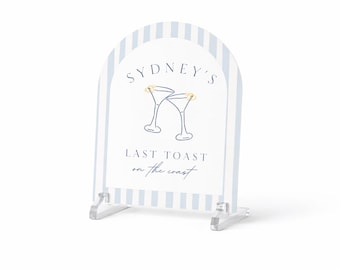 May include: Clear acrylic sign with a blue and white striped border. The sign features the text "Sydney's Last Toast on the Coast" and a graphic of two martini glasses with gold accents. The sign is supported by clear acrylic feet.