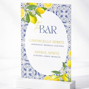 May include: A rectangular bar menu sign featuring a blue and white tile pattern border with yellow lemon illustrations. The center oval displays the text The BAR, LIMONCELLO SPRITZ with ingredients, and APEROL SPRITZ with ingredients. The sign stands on a white base.