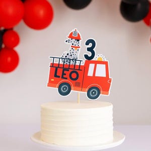 May include: A white iced cake on a white cake stand, decorated with a red fire engine cake topper. The topper shows a Dalmatian dog in a firefighter's hat and the number 3. Black and red balloons are in the background.