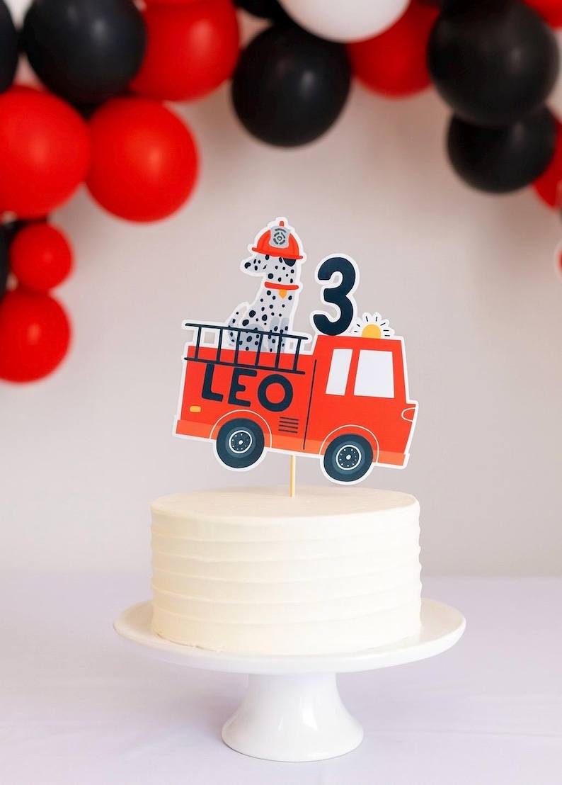 May include: A white iced cake on a white cake stand, decorated with a red fire engine cake topper. The topper shows a Dalmatian dog in a firefighter's hat and the number 3. Black and red balloons are in the background.