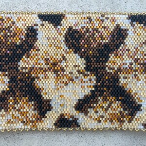 May include: A rectangular beaded cuff bracelet featuring a repeating geometric pattern in shades of black, dark brown, cream, and metallic gold. The design is composed of small, uniform glass beads arranged in a dense, woven grid structure.