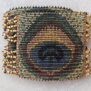 May include: A rectangular beaded bracelet with a shimmering, iridescent pattern. The bracelet features a mosaic of small beads in shades of green, gold, and brown. The edges are finished with a decorative fringe of gold beads and dark blue accents.