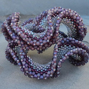 May include: A purple and silver beaded sculpture with a complex, interwoven design. The sculpture is made of small, round beads that are arranged in a series of loops and curves.