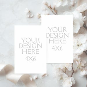 May include: Two white 4x6 inch cards with the text "YOUR DESIGN HERE" are displayed. The cards are surrounded by white flowers and a cream-colored ribbon on a marble background.