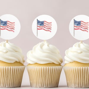 May include: Three cupcakes with white frosting topped with round paper circles featuring a watercolor American flag design.