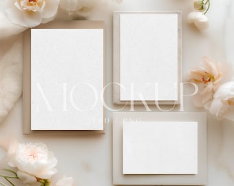 May include: A mockup featuring three blank white cards in different sizes, arranged on a marble surface. The cards are set within light gray frames, with a beige envelope. Soft white and peach flowers are scattered around the cards, creating a delicate, elegant composition.