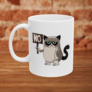 Puede incluir: Taza de café de cerámica blanca con una imagen de dibujos animados de un gato gruñón sosteniendo un cartel que dice "NO".