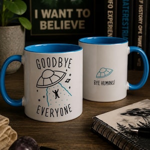 Op de afbeelding: Twee witte keramische mokken met blauwe binnenkant en handvatten. Eén mok heeft een zwarte en blauwgroene afbeelding van een UFO die een stokfiguur ontvoert met de tekst "GOODBYE EVERYONE." De andere mok heeft een UFO-afbeelding en de tekst "BYE HUMANS!"