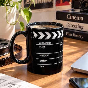 Puede incluir: Una taza de cerámica negra con un gráfico de claqueta de cine en blanco. El diseño incluye el texto PRODUCTION, SCENE, TAKE, DIRECTOR, CAMERA y DATE con líneas para escribir. La taza tiene un asa curva y contiene una bebida oscura.