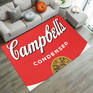 Puede incluir: Una alfombra rectangular roja con el diseño de la etiqueta de sopa Campbell's Condensed. La alfombra presenta el logotipo de Campbell's en escritura blanca, la palabra "CONDENSED" en blanco y un sello dorado. La alfombra está sobre un suelo de madera.