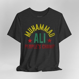 May include: Dark gray t-shirt with the text "MUHAMMAD ALI" in yellow, "ALI" in green, and "PEOPLE'S CHAMP" in red. Two red stars flank the word "ALI". The shirt has a crew neck and short sleeves.