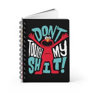 May include: A spiral-bound notebook with a black cover featuring a red cartoon character with outstretched arms. The text "DON'T TOUCH MY SHIT!" is written in white and teal. The notebook has a white interior and a black spiral binding.
