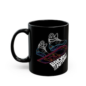 Puede incluir: Taza de cerámica negra con un gráfico de neón de zapatillas futuristas y una tabla voladora. Las palabras "BACK TO THE FUTURE" están impresas en blanco. La taza tiene un asa negra y un acabado brillante.