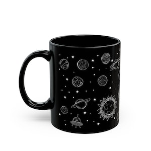 Puede incluir: Taza de cerámica negra con una ilustración blanca de planetas, estrellas y un sol. La taza tiene un asa curva y una forma cilíndrica. El diseño incluye Saturno, un OVNI y otros cuerpos celestes.