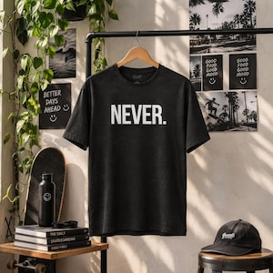 Puede incluir: Una camiseta de manga corta gris carbón con la palabra NEVER. impresa en letras mayúsculas blancas grandes en el pecho. La prenda se muestra en una percha de madera contra una pared clara con varios objetos decorativos cerca.