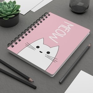 May include: A pink spiral-bound notebook with the word "MEOW" in white and a white cat illustration. The notebook is open, revealing blank pages. Two black pencils and a small succulent plant in a black pot are also visible.