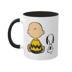 May include: White ceramic mug with a black interior and handle. The mug features a cartoon illustration of Charlie Brown and Snoopy. Charlie Brown is wearing his signature yellow shirt with a zig-zag pattern and is sitting with his back to the viewer. Snoopy is standing next to him, looking to the right.
