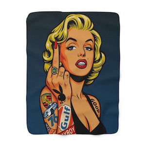 Può includere: Una morbida coperta con un'illustrazione in stile pop art di una donna con capelli biondi, rossetto rosso e tatuaggi. La donna sta facendo il dito medio. La coperta ha uno sfondo blu scuro.