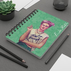 Puede incluir: Cuaderno de espiral con una ilustración de Frida Kahlo con una corona de flores y una camiseta blanca con las palabras "daft punk". El cuaderno tiene un fondo de patrón floral verde y las palabras "Frida Kahlo" en escritura blanca.