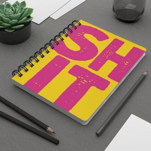 Puede incluir: Un cuaderno de espiral con una cubierta de color amarillo brillante que presenta el texto grande, en negrita y desgastado de color rosa que dice "ISH IT". El cuaderno tiene una encuadernación de alambre negro en el lado izquierdo y está sobre una superficie gris oscuro.