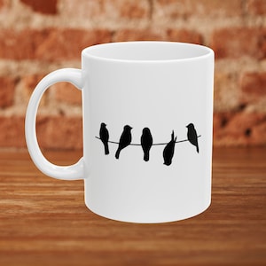 Puede incluir: Taza de café de cerámica blanca con una silueta negra de cinco pájaros posados ​​en un alambre.