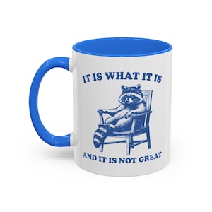 May include: A white ceramic mug with a bright blue handle and rim. The front features a blue graphic of a raccoon sitting in a wooden chair. Above the raccoon is the text IT IS WHAT IT IS and below is the text AND IT IS NOT GREAT.