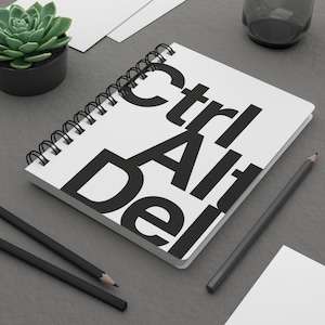 Puede incluir: Un cuaderno blanco con espiral con las palabras "Ctrl Alt Del" impresas en letras negras grandes. El cuaderno está sobre una superficie gris, junto a dos lápices negros, una pequeña planta suculenta en una maceta negra y un vaso transparente.