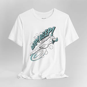 Puede incluir: Camiseta blanca con un personaje de dibujos animados de un pájaro en movimiento y las palabras "Beep! Beep!" en una fuente estilizada. El diseño utiliza una combinación de colores azul y blanco, con líneas de movimiento y un elemento circular.