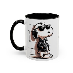 Puede incluir: Taza de cerámica blanca con interior y asa negros. La taza presenta una ilustración de dibujos animados de un perro con gafas de sol y una chaqueta de cuero negra. El perro está apoyado en una pared de ladrillos.