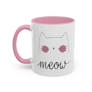 Puede incluir: Una taza de cerámica blanca con interior y asa de color rosa. La taza presenta un dibujo de línea negra de un gato con círculos rosas para los ojos y la palabra "miau" escrita debajo del gato.