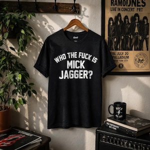 Puede incluir: Una camiseta de manga corta en color gris carbón con el texto blanco "WHO THE FUCK IS MICK JAGGER?" impreso en el pecho. La prenda tiene un cuello redondo y se muestra colgada en una percha de madera sobre una pared clara.