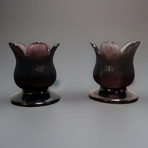 May include: Two matching purple glass candle holders with a tulip-shaped design. Each piece features a ribbed, textured exterior surface, a wide circular base, and a scalloped rim that mimics the appearance of flower petals in deep violet glass.
