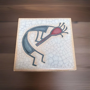 May include: A square ceramic tile featuring a crackled off-white glaze finish. The center displays a dark gray stylized figure of a flute player with a prominent red head and hair-like extensions, rendered in a flat, graphic, and minimalist style.