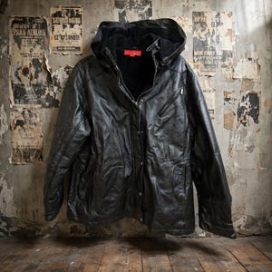May include: A black leather jacket featuring a hood, a front zipper closure, and a snap button placket. The jacket includes a diagonal zippered chest pocket and a red interior label near the collar, showcasing a textured finish throughout.