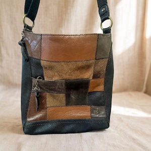 May include: A crossbody bag featuring a patchwork design of brown, tan, and black leather panels. The bag includes a front zippered pocket with a tassel pull, a black fabric shoulder strap attached with brass-tone rings, and the text the sak.