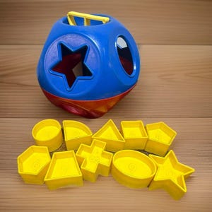 May include: A blue and red plastic shape-sorting toy with a yellow handle on top. Ten yellow plastic shapes are arranged in front, each featuring a different geometric form and a raised number from one to ten embossed on the surface.