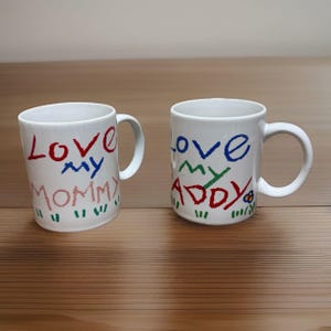 May include: Two white ceramic mugs with handwritten text. One mug says "Love my Mommy" in red, blue, and pink. The other says "Love my Addy" in blue, green, and red. Both mugs have a simple handle.