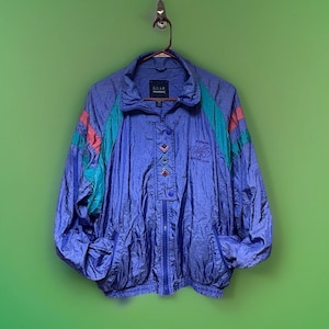 May include: A blue windbreaker jacket featuring a high collar, a partial front zipper, and snap buttons. The jacket includes teal and pink color-blocked accents on the shoulders and sleeves, with embroidered geometric patterns and text on the front.