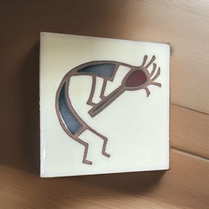 May include: A square ceramic tile with a glossy off-white finish featuring a stylized brown line drawing of a flute player. The figure has a dark red head and dark blue body segments, depicted in a hunched posture with thin limbs and hair.