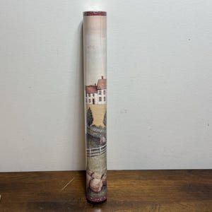 May include: A vertical roll of wallpaper featuring a repeating pastoral scene with a white house, a red roof, green trees, and a white fence set against a beige background. The top and bottom edges of the roll have a thin, dark red decorative border.