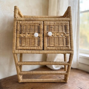 May include: A light brown woven wicker wall shelf featuring two small cabinet doors with round white knobs. The unit includes an open top section and two horizontal bars at the bottom, constructed from natural rattan material with a woven texture.