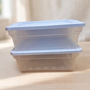 May include: Two rectangular, clear plastic storage containers stacked vertically. Each container features a light blue, snap-on lid with a flat top and slightly raised edges. The transparent base of each container reveals a smooth interior.