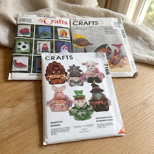 May include: Three craft pattern envelopes featuring illustrations of various projects. One envelope shows small fabric baskets shaped like animals and characters, with text stating baskets are 13.5 inches high. Other envelopes display wall hangings and fish designs.