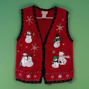 May include: A red V-neck knit vest featuring embroidered snowmen and white snowflake patterns. The garment has a black trim with a cross-stitch design along the edges and a front button closure. A label reads Basic Editions Holiday.
