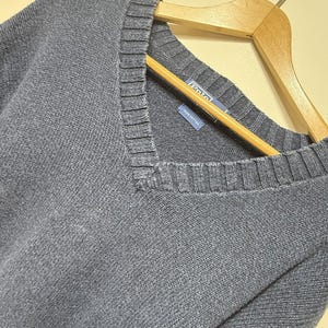 May include: A blue V-neck sweater made of 100% cotton. The garment features a ribbed neckline and a blue interior label with the text Polo Ralph Lauren and 100% cotton printed in white. The sweater is displayed on a wooden hanger.