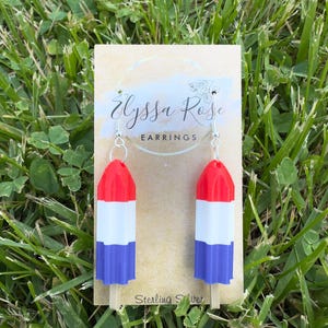 Può includere: Orecchini a forma di ghiacciolo rosso, bianco e blu, con ganci in argento sterling. Gli orecchini sono presentati su una carta color crema con il testo "Elyssa Rose Earrings" e "Sterling Silver". Sfondo di erba verde.