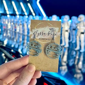 May include: Light blue Death Star earrings on a brown card with the text "Elyssa Rose Earrings" and "Sterling Silver". The earrings are held in front of a blue background with white figures.