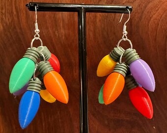May include: A pair of earrings featuring colorful, 3D-printed light bulbs. Each earring has a cluster of bulbs in various colors, including green, blue, orange, yellow, red, and purple, with silver-colored hooks.